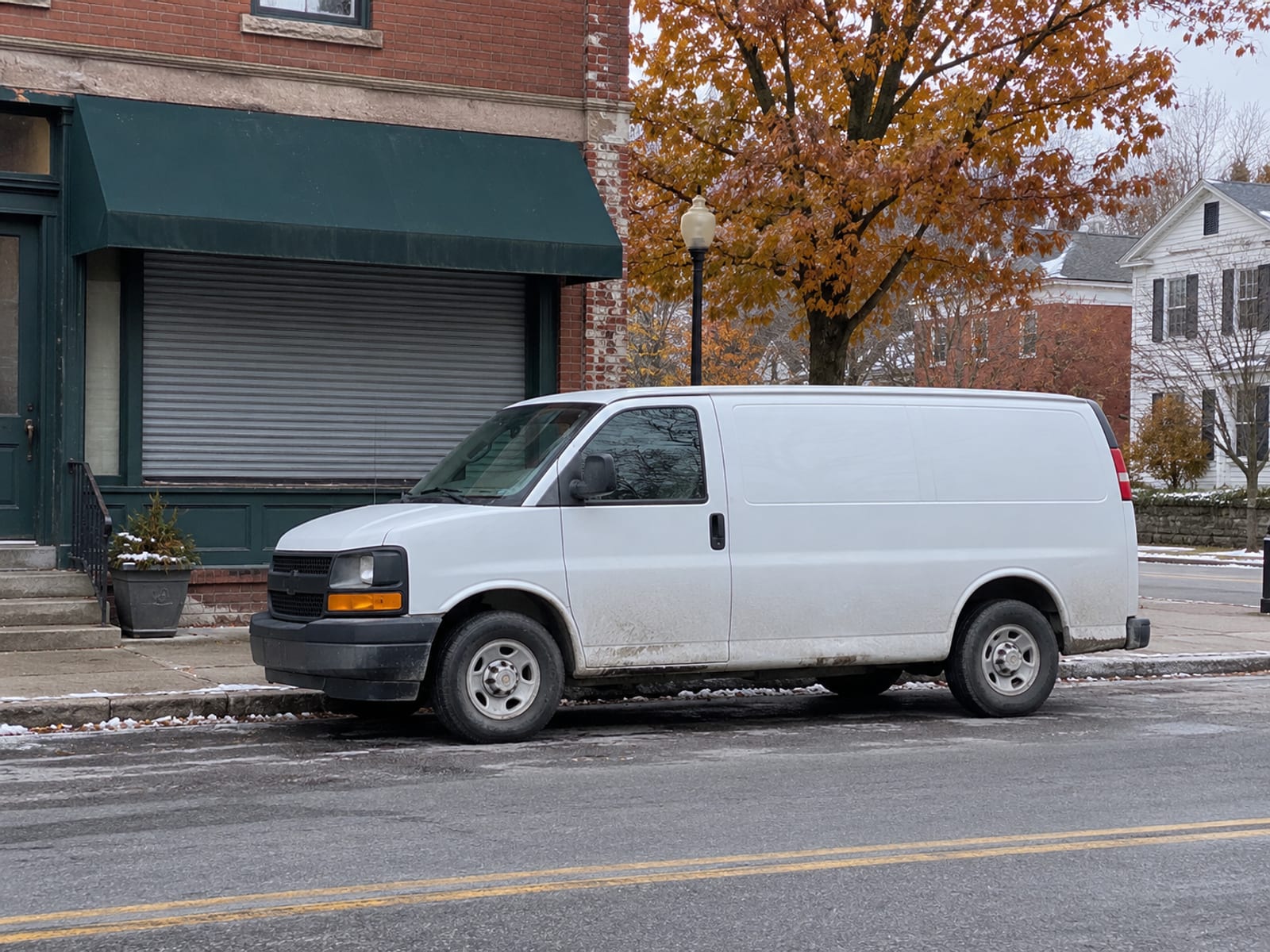 A plain white work van parked in front of a small brick storefront on a village street