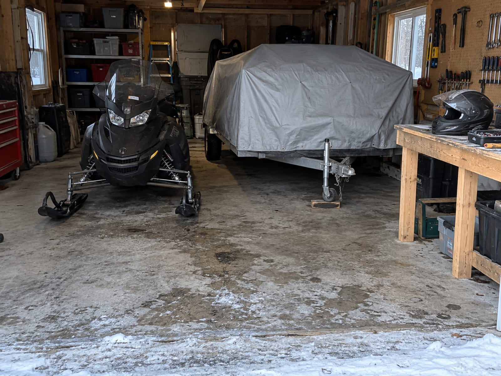 A snowmobile in a garage beside a covered trailer and a helmet on a workbench