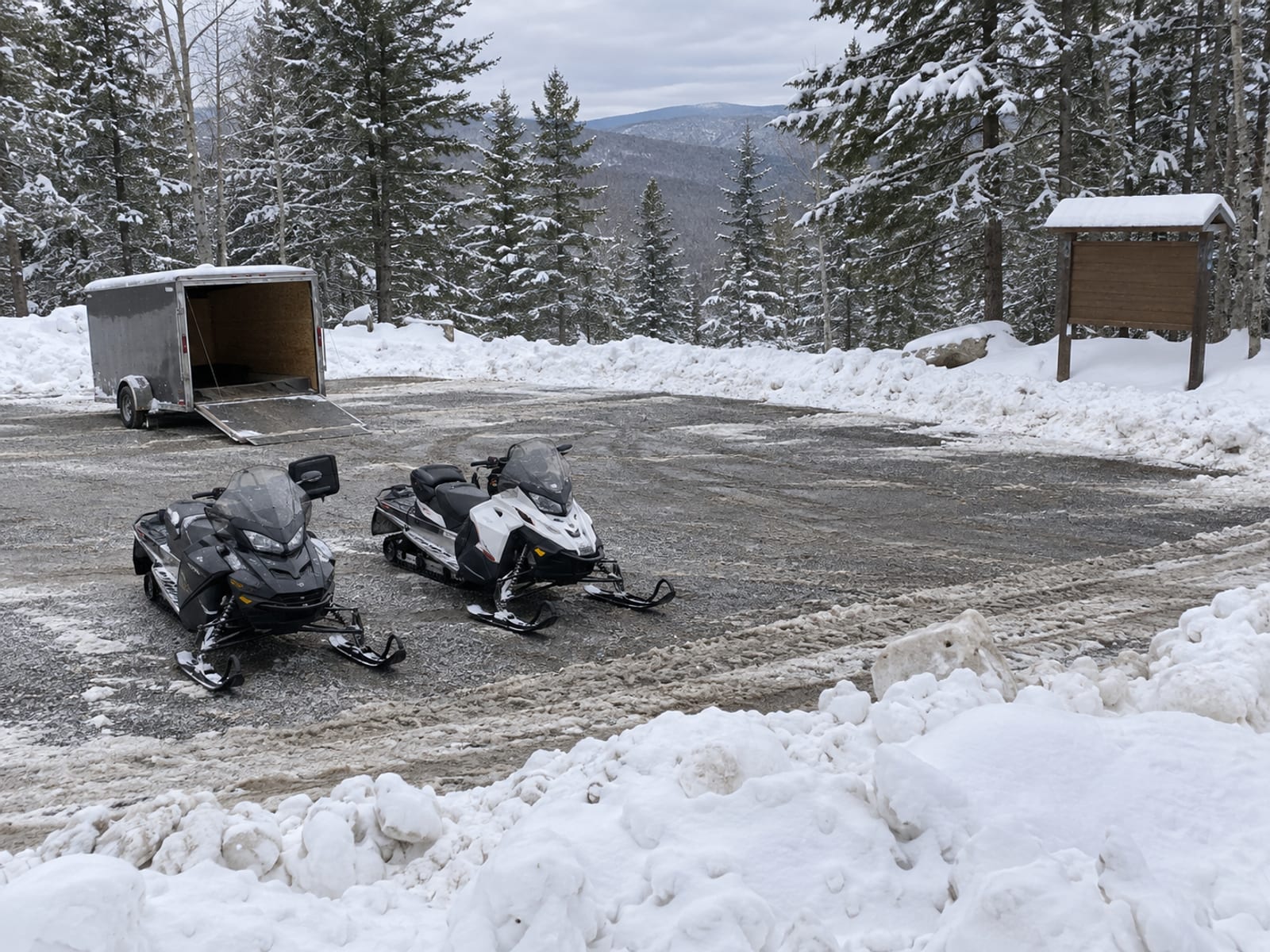 A snowy trailhead parking area with a trailer and two parked snowmobiles beside pine trees
