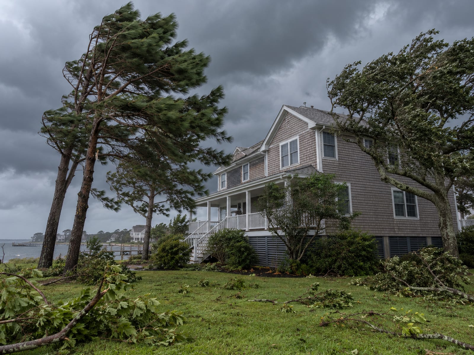 Wind-bent trees and scattered branches beside a coastal house under dark storm clouds