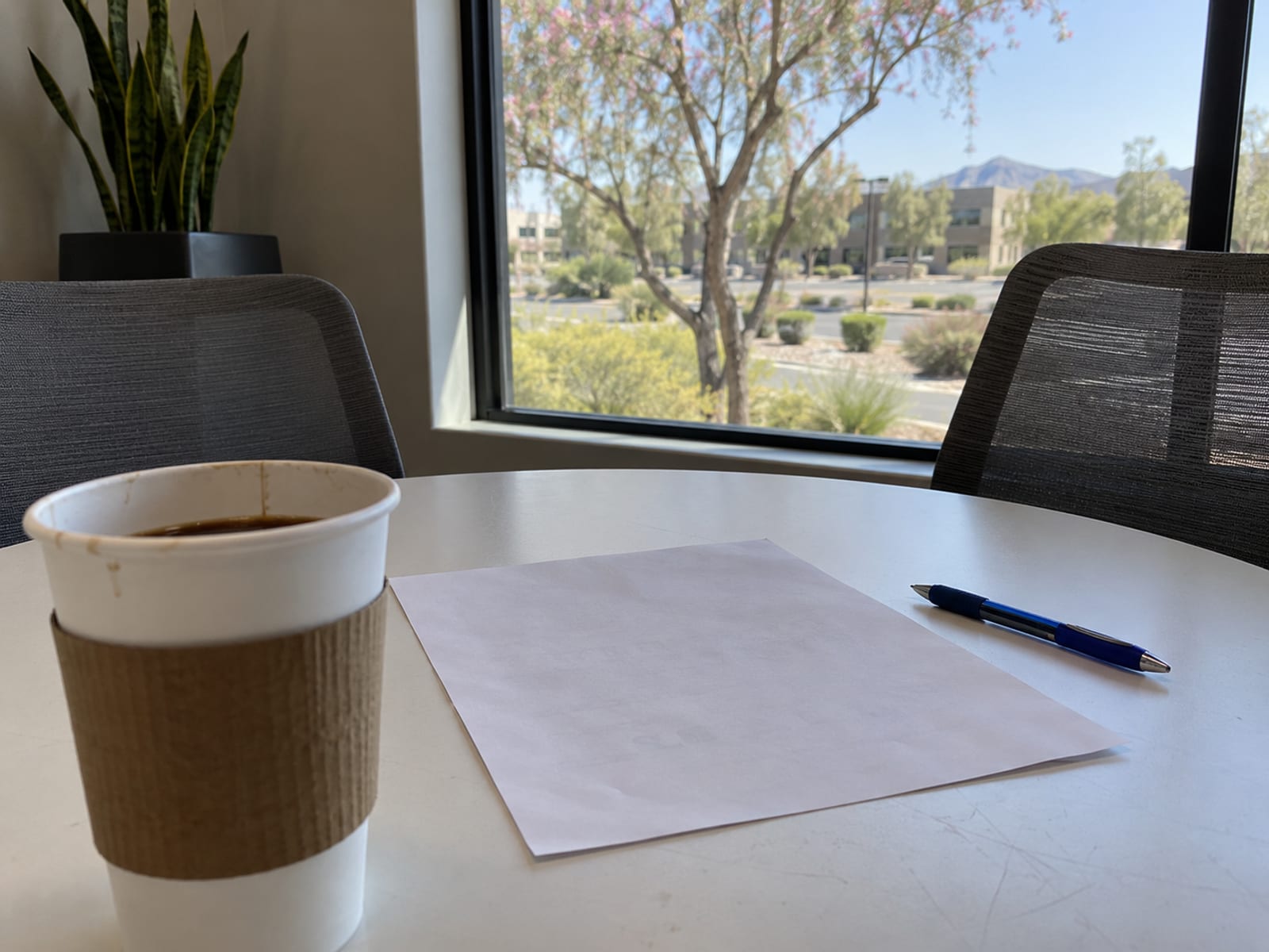 An office table with a printed worksheet face down, a pen and a cup of coffee by a window