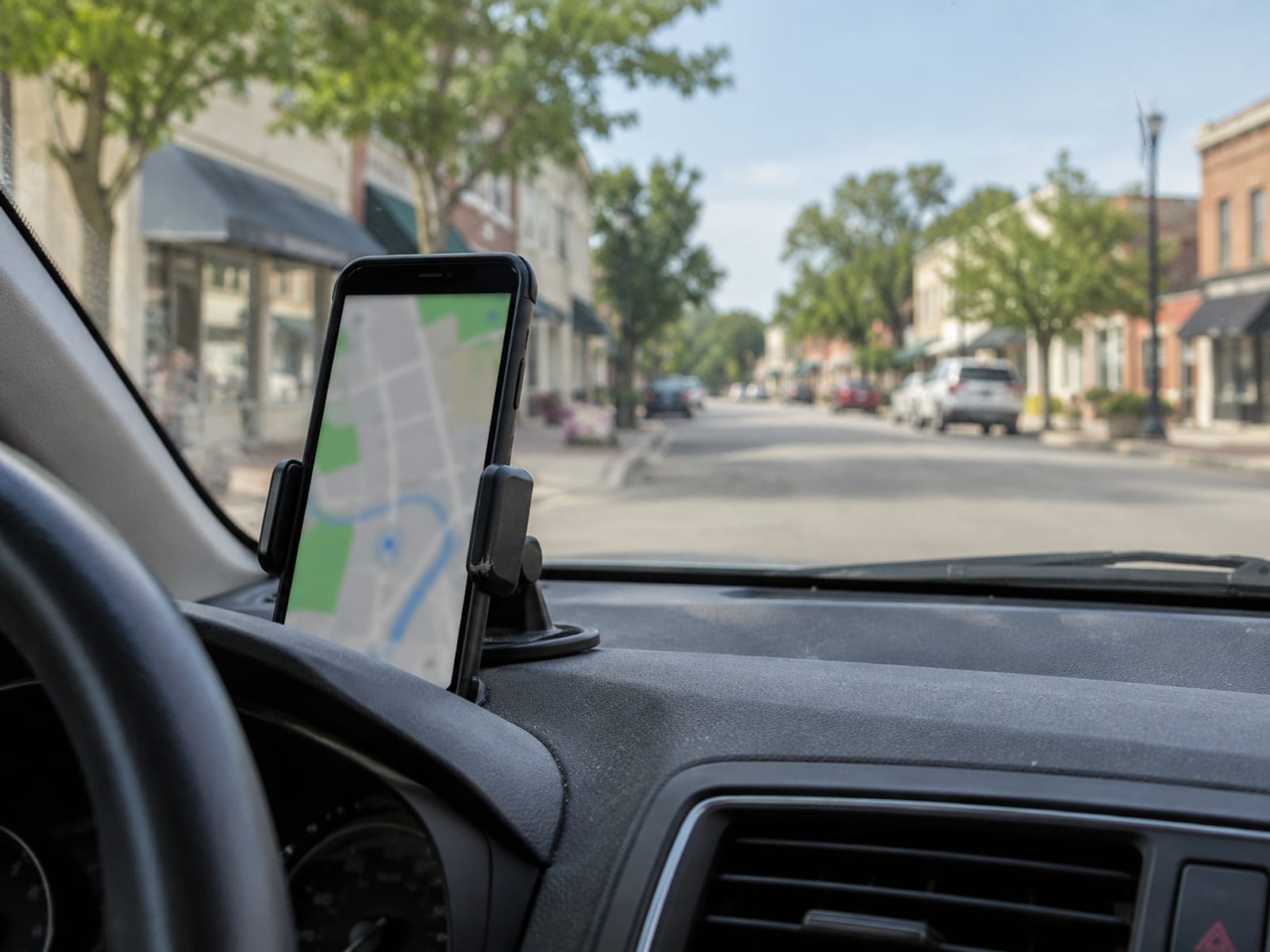 A phone with a blurred map screen on a car dashboard mount, a small-town street beyond
